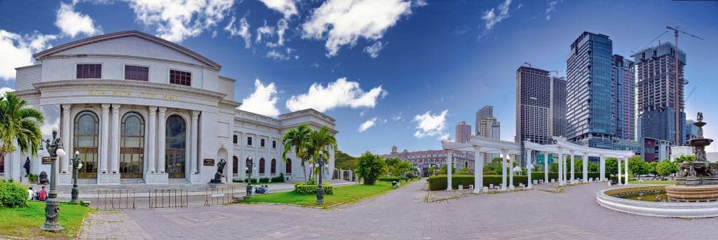 Panoramic photo of Diamond City Hall in Phnom Penh, Cambodia, and the surrounding urban landscape. The sky is blue and the grass and plants are green. We can guess that a site is under construction because there are many cranes there.