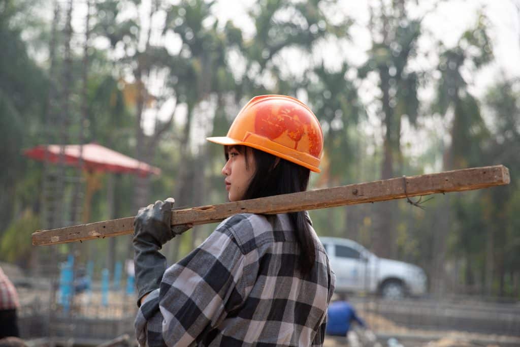A young Cambodian woman wearing an orange hard hat is wearing a grey checked shirt and carrying a wooden beam on her shoulder.
