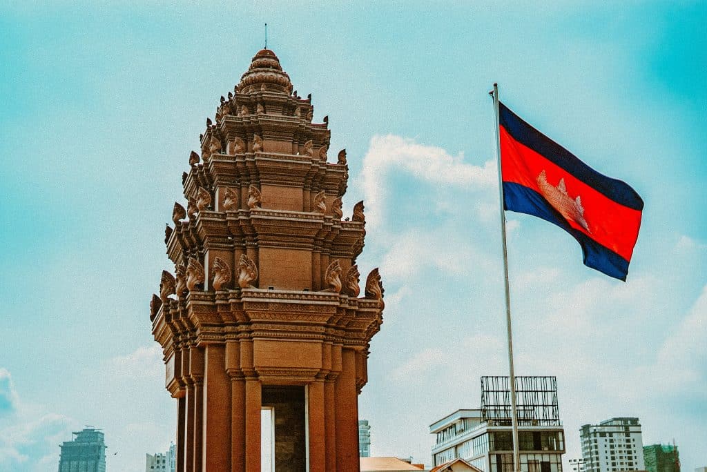 The Cambodian flag is raised and flying in the wind next to a traditional monument in Cambodia, in Phnom Penh, under a clear blue sky.