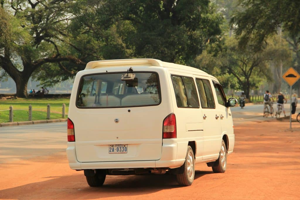 Photo taken from the back of a white van travelling on the orange sand of a road in Krong Siem Reap, Cambodia.
