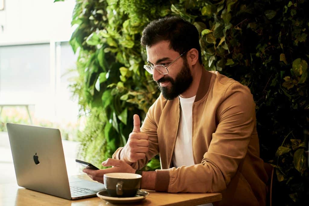 A bearded brown-haired man wearing glasses and a brown beret is sitting at a café table and working on his laptop via video call. He is smiling, holding his phone in one hand and giving a thumbs up with the other. There is a green wall behind him.