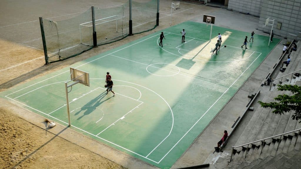 Aerial photo of a school basketball court in Seoul, South Korea. A group of boys are playing basketball. The court is green and the entire surrounding area is made up of sand and concrete.