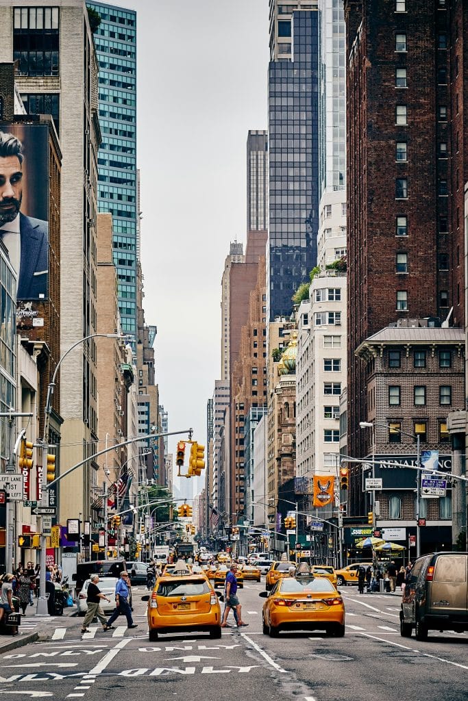 People crossing a pedestrian crossing in New York City. The famous yellow taxis are stopped at the pedestrian crossing. Large advertising posters adorn the city's buildings.