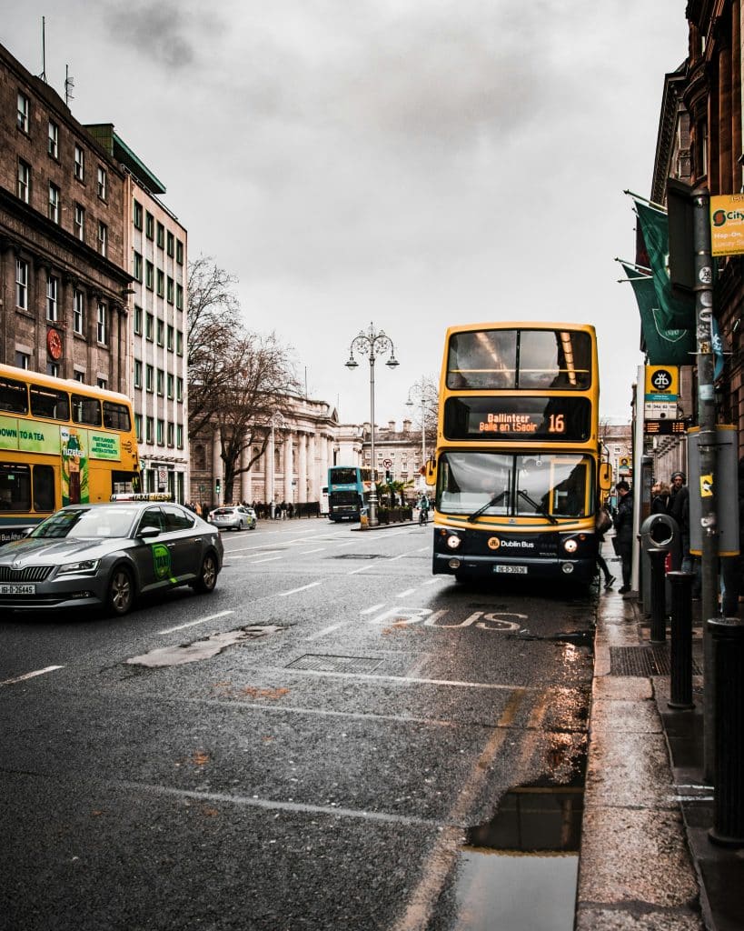 A yellow double-decker bus stops at a stop sign in a street in Dublin, Ireland. The weather is gloomy and it has rained, as evidenced by the puddles. A black car passes the bus in the next lane.