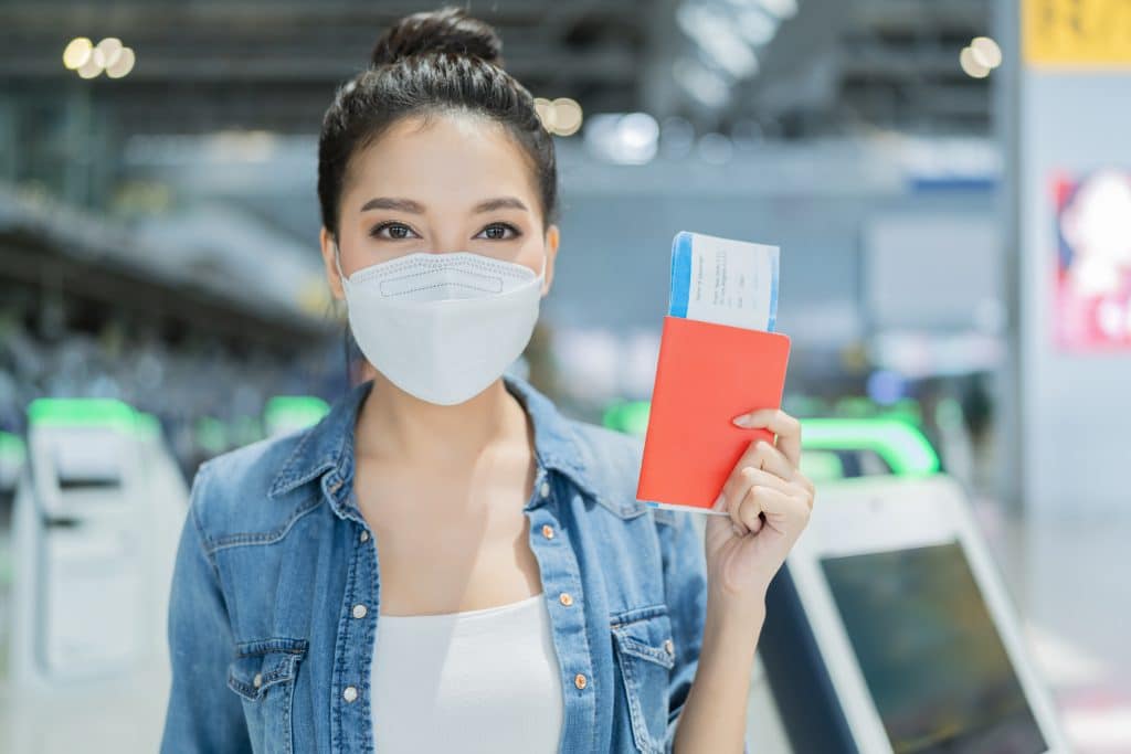 An Asian woman wearing a mask, a white T-shirt and a denim jacket is holding a passport and an airline ticket in her hand. She is in an airport and appears to be smiling behind her mask.