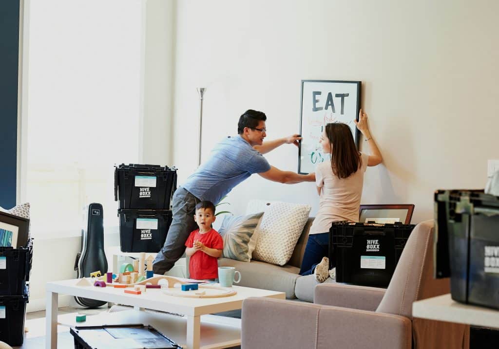 Two parents hang a picture in the new living room where they have just moved in with their little boy. Wooden toys are placed on the coffee table while the parents kneel on the sofa to hang the picture.