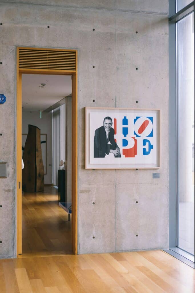 A poster frame with a contemporary-style photo of Obama hangs on the wall of a light grey concrete office room, to the left of a bay window and to the right of a door with a mustard yellow frame. It all adds up to a modern, industrial feel.