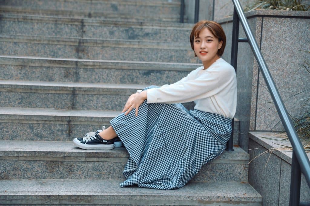 A young woman with short hair of Korean origin is sitting on the steps of an outdoor staircase and smiling at the camera. She is wearing a long skirt, trainers and a white sweatshirt.