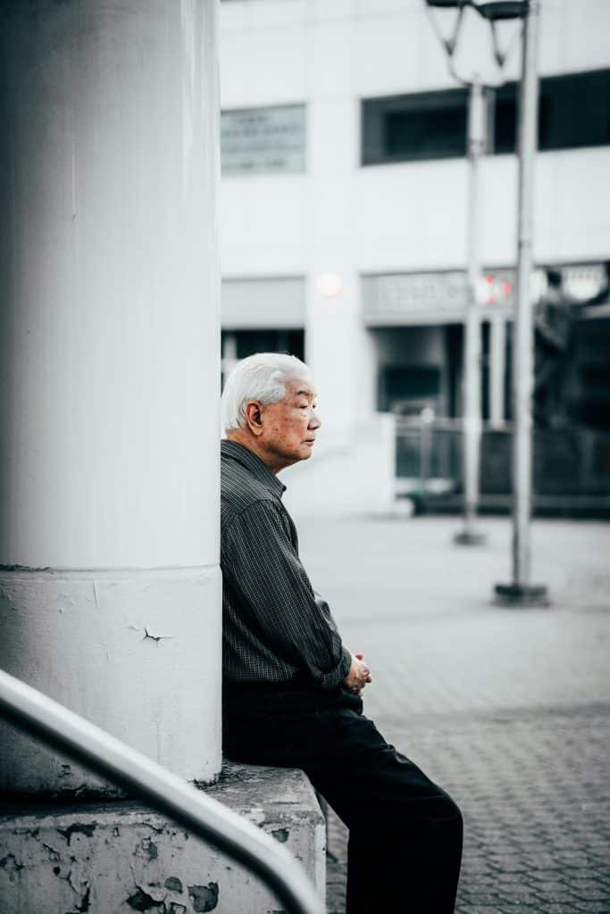 An old Asian man is sitting at the foot of a stone column in the street, holding his hands and gazing at the surrounding landscape.
