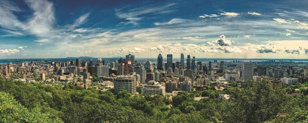 Aerial photo of Montreal in the Quebec region of Canada. The foreground of the photo shows a large expanse of greenery and forest. The background shows the city and its tall buildings.