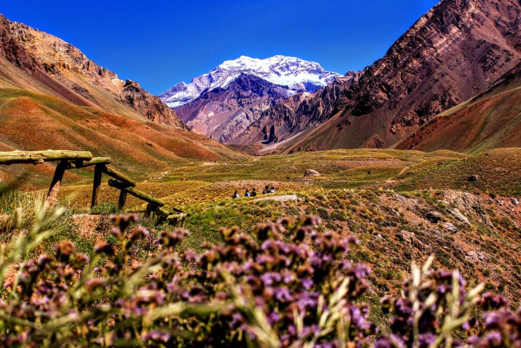 A group of people are hiking in a magnificent green plain surrounded by snow-capped mountains. They descend along a fenced wooden path. The weather is very sunny.