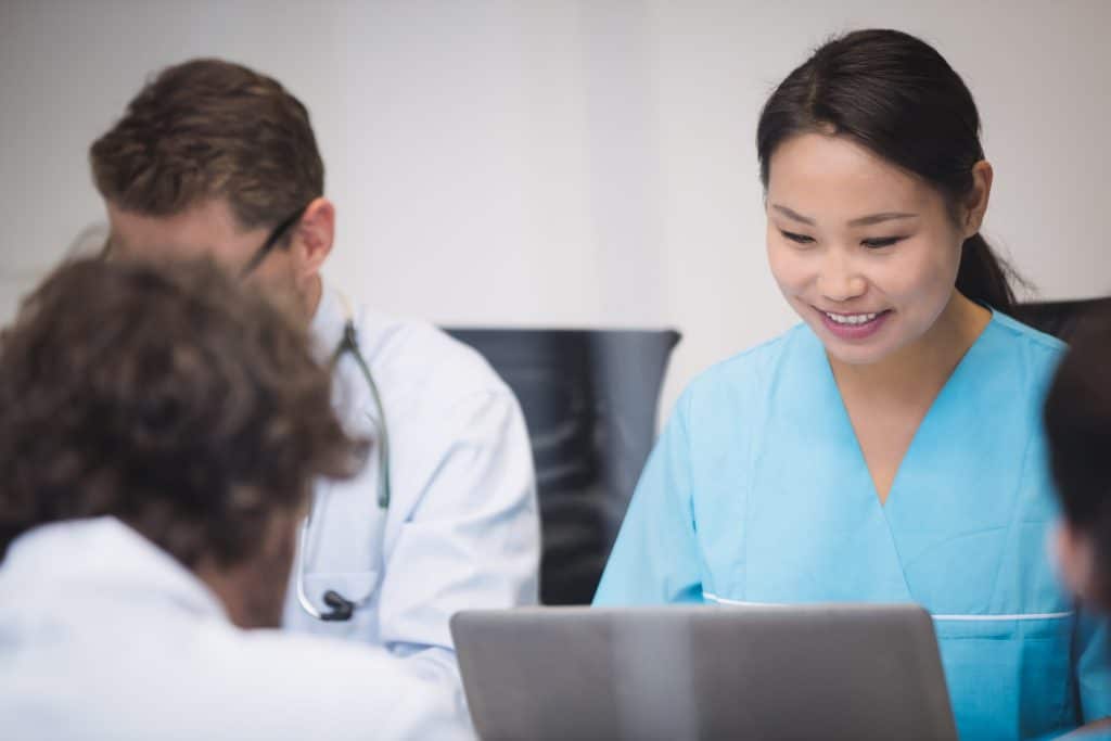 Medical personnel are gathered around a table. A Japanese nurse smiles as she consults her laptop. Two male colleagues are seated beside her.