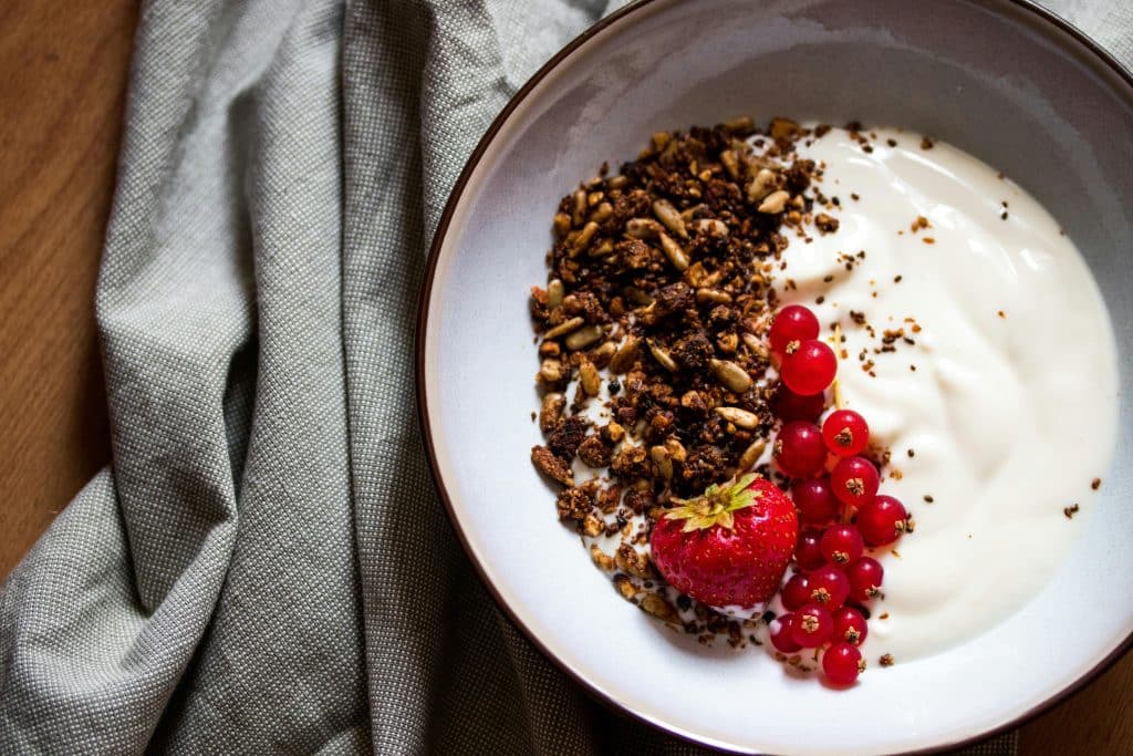 A bowl of yoghurt with granola, a strawberry and redcurrants, placed on a wooden table with a grey napkin.