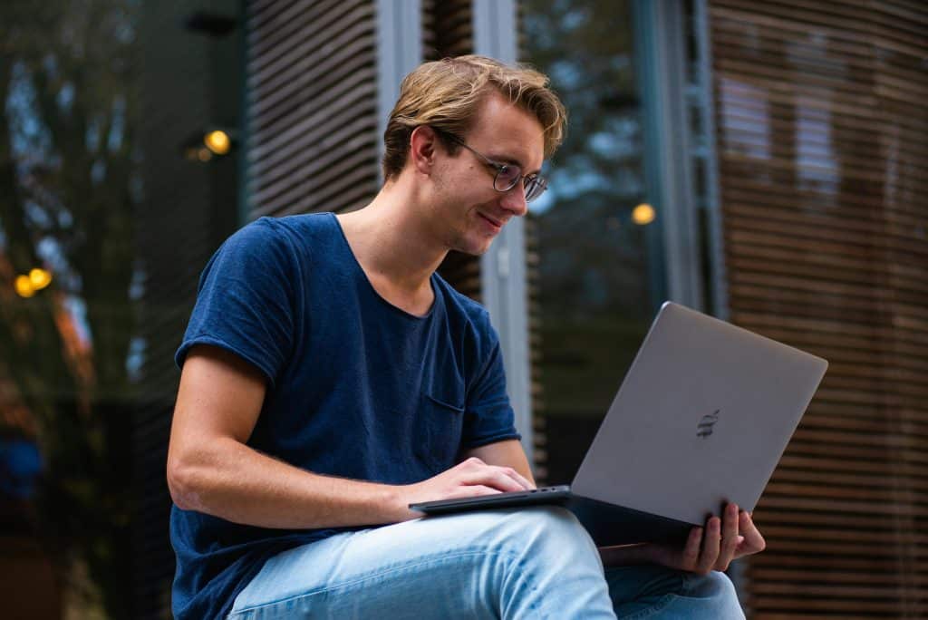 A young man with glasses is working, smiling, on his laptop computer, which is resting on his knees. He is wearing a dark blue T-shirt and jeans.