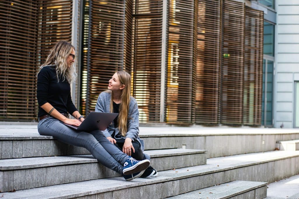Photo of two students sitting on the stairs of a university in the Netherlands. One of the women is wearing glasses and is holding her laptop on her legs. The other student is looking at her and smiling.
