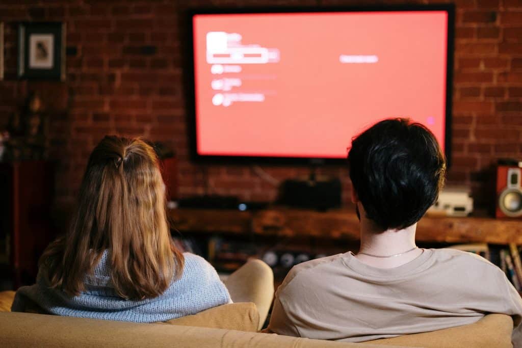 A couple seen from behind are watching a film on television, sitting on a sofa.
