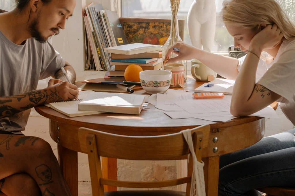 A man and a woman in their twenties are studying at a wooden table in a dining room. The man is copying notes and the young woman is reading notes while serving a cup of tea.