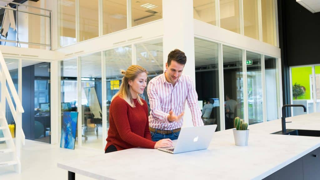 A man and a woman are work colleagues and are chatting together in their startup-style workplace. They are both looking at a laptop. The background of the photo shows an office room with large bay windows.