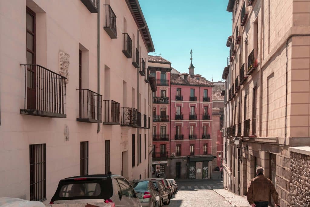 A small street in Madrid, Spain. Cars are parked along the pavement and there are small pink buildings. An old man is walking down the street.