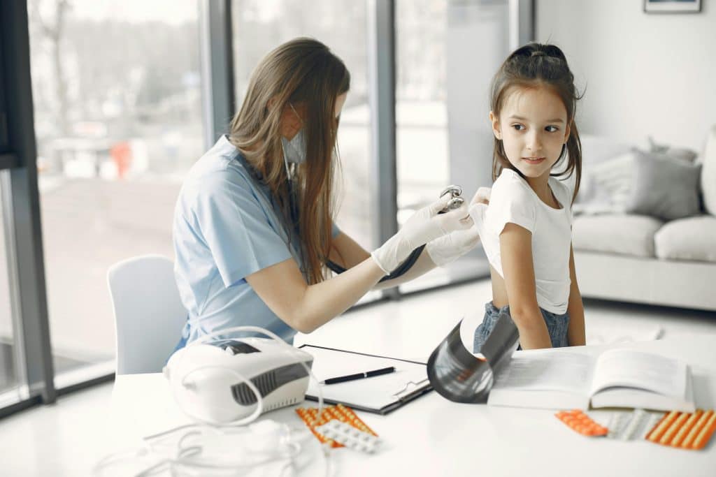 A nurse examines a little girl in her home. She listens to the girl's breathing by placing a stethoscope on her back. A device and documents are scattered on the table.
