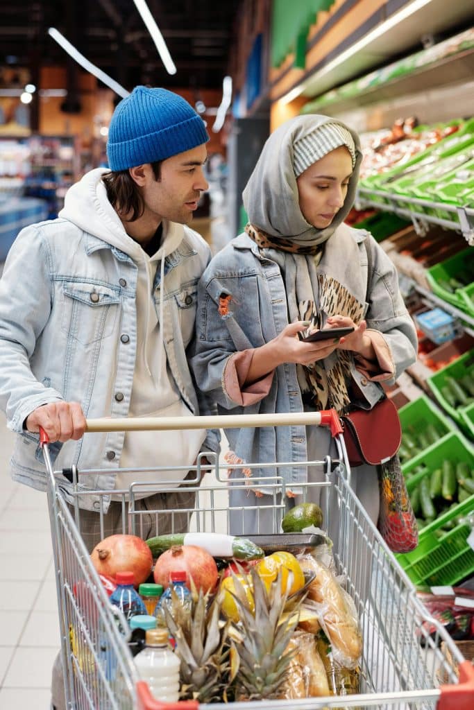 A couple is shopping in a supermarket. Their trolley is full of products. The woman is calculating their spending on her phone.