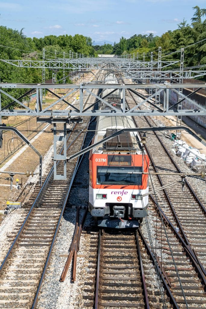 Photo of a train travelling on the rails near the Spanish capital. The train is white and orange.