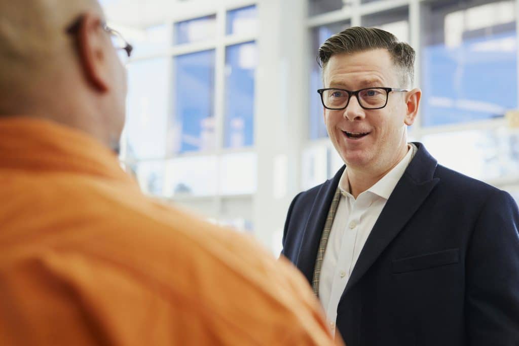 A man in a suit and glasses is talking to a colleague. The photo is taken over the colleague's shoulder.