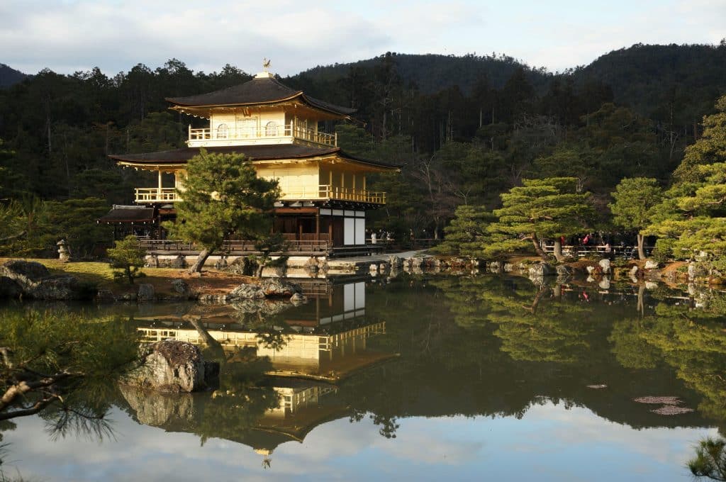 A Japanese temple by a lake and surrounded by a forest. The landscape is reflected in the water. The photo was taken at sunrise or sunset.