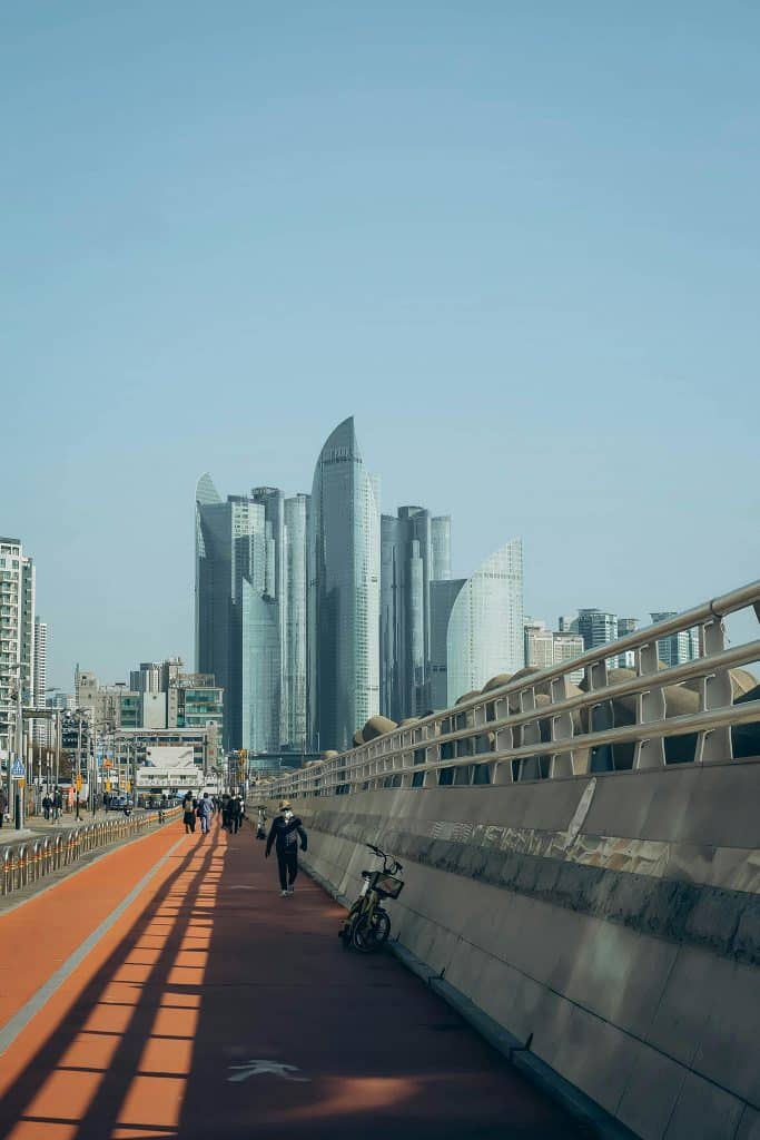 Residents walk on a long bridge in Busan, South Korea. The photo was taken on a sunny day. Large modern towers are in the background of the photo.