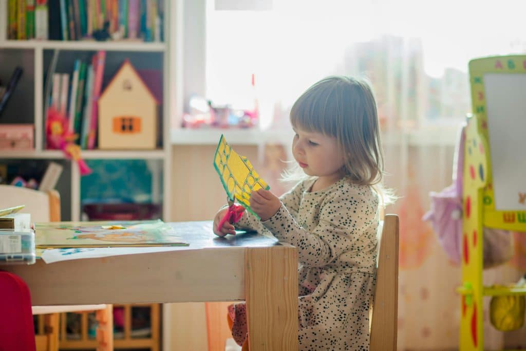 A little blonde girl is doing some cutting out in nursery school. She is sitting on a wooden chair, with colouring books and other books on the wooden table and a children's blackboard behind her. The bookcase in the background contains books and a wooden playhouse.