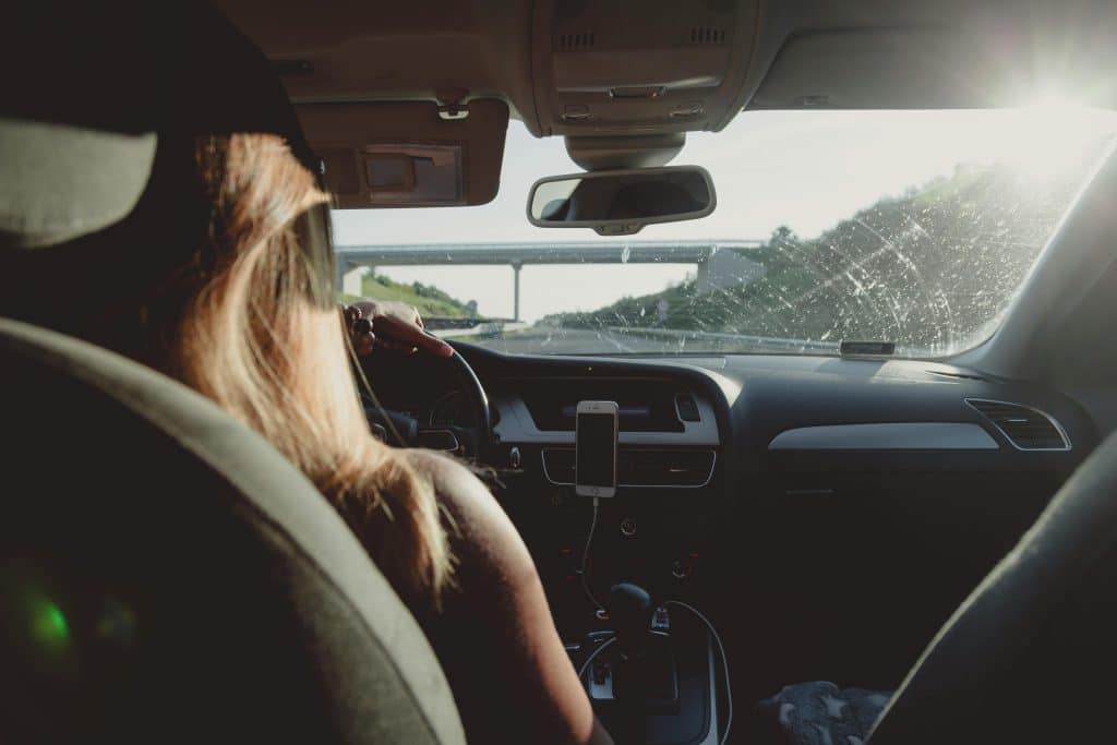 Photo taken from the back seat of a car showing a woman at the wheel, following the directions of her GPS on her phone, attached to the centre console of the car. The car is travelling on a motorway. The windscreen has not been cleaned properly.