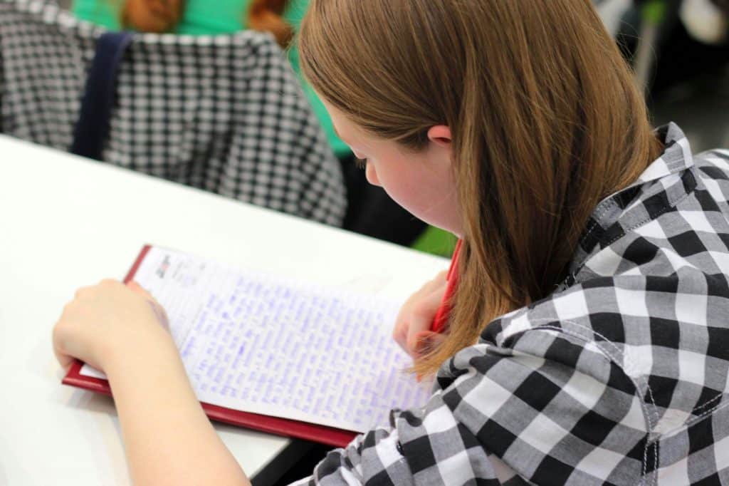 A teenage pupil is taking notes in her exercise book in class. She is wearing a black and white checked shirt.