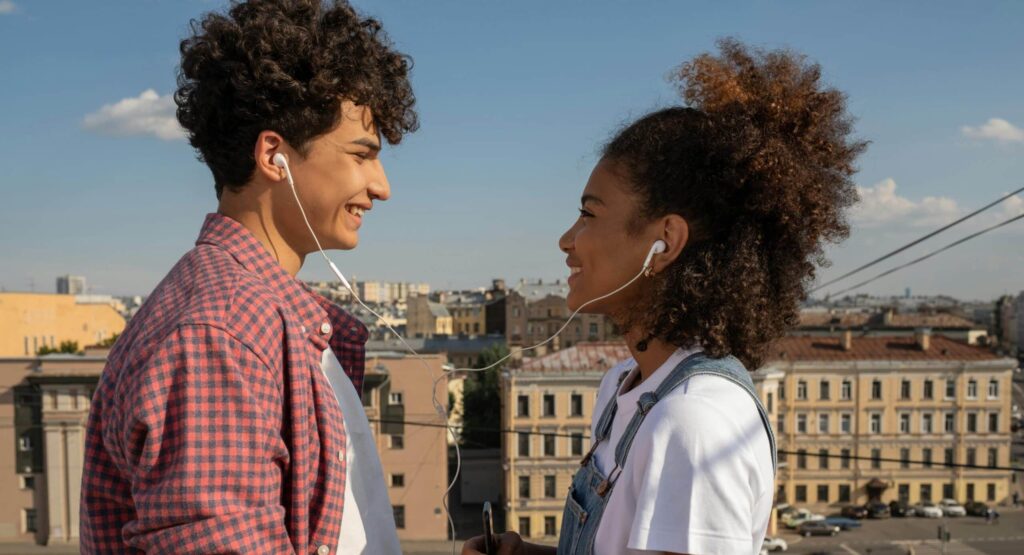Two young people, a man and a woman, smile at each other and share the same headphones, listening to music on a phone held by the woman. The young man is wearing a red checked shirt and the young woman a white T-shirt with denim overalls. They are standing on an elevated terrace overlooking a city-centre avenue.