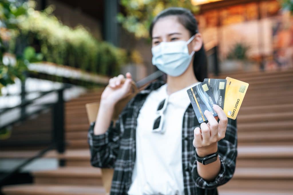 An Asian woman wearing a mask is holding three credit cards in her hand. She is carrying a handbag on her shoulder and wearing sunglasses on the collar of her T-shirt. Her hair is tied back in a ponytail.