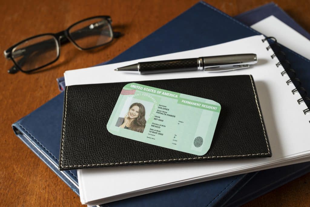 Top view of an American green card placed on a wallet and folders next to a beautiful pen and black glasses, on a wooden table.