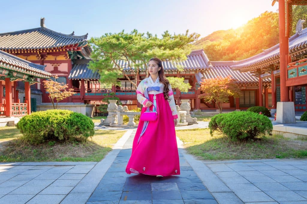 A young woman walks through a traditional Korean garden in a hanbok. She is also carrying a small pink handbag.